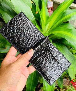 Hình ảnh ví kẹp tiền da cá sấu đen VOA5, phong cách thời trang, được cầm trên tay giữa cảnh lá xanh tươi.