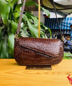 Hình ảnh túi đeo vai da cá sấu HM-20 màu nâu, đặt trên bàn gỗ, thể hiện phong cách thời thượng giữa cây xanh.
