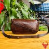Hình ảnh túi đeo vai nữ cao cấp da cá sấu HM-20 màu nâu, đặt trên bàn gỗ giữa cây xanh và chợ, nổi bật phong cách sang trọng.