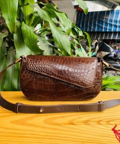 Hình ảnh túi đeo vai nữ cao cấp da cá sấu HM-20 màu nâu, đặt trên bàn gỗ giữa cây xanh và chợ, nổi bật phong cách sang trọng.