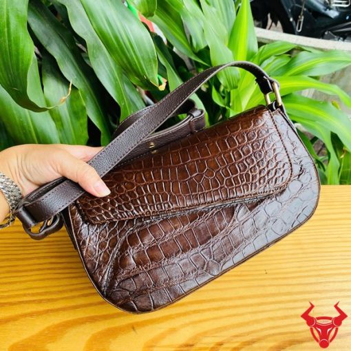 Hình ảnh túi đeo vai da cá sấu HM-20 màu nâu, đặt trên bàn gỗ, nổi bật giữa cây xanh với phong cách thời trang.
