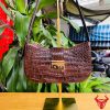 Hình ảnh túi đeo vai nữ da bụng cá sấu HM-19 màu nâu, khóa vàng, đặt trên giá gỗ giữa cây xanh và chợ, nổi bật phong cách sang trọng.