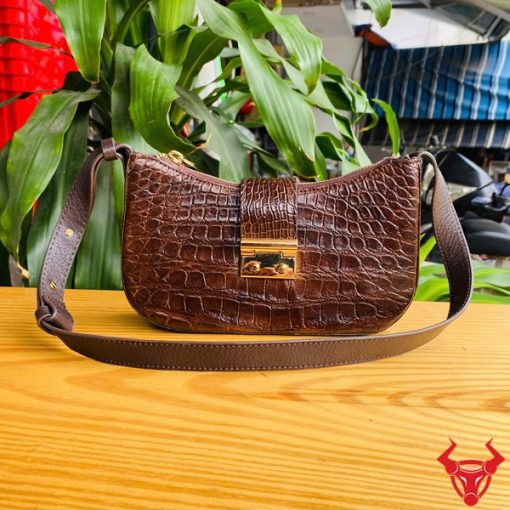 Hình ảnh túi đeo vai da bụng cá sấu HM-19 màu nâu, khóa vàng, đặt trên giá gỗ, thể hiện phong cách thời thượng giữa cây xanh.