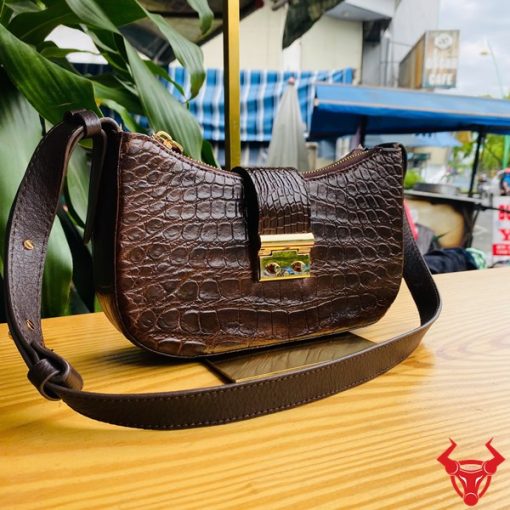 Hình ảnh túi đeo vai da bụng cá sấu HM-19 màu nâu, khóa vàng, đặt trên giá gỗ, nổi bật với phong cách sang trọng.