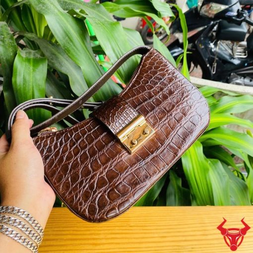 Hình ảnh túi đeo vai da bụng cá sấu HM-19 màu nâu, khóa vàng, đặt trên giá gỗ, thể hiện phong cách sang trọng và cá tính.