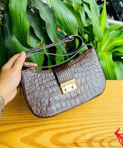 Hình ảnh túi đeo vai da bụng cá sấu HM-19 màu nâu, khóa vàng, đặt trên giá gỗ, nổi bật giữa cây xanh với phong cách thời trang.