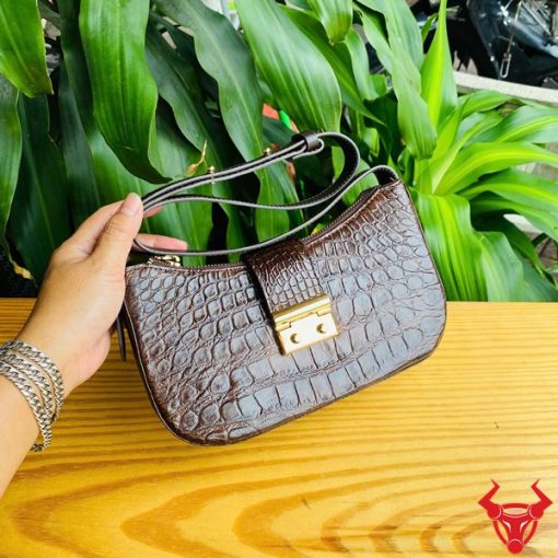 Hình ảnh túi đeo vai da bụng cá sấu HM-19 màu nâu, khóa vàng, đặt trên giá gỗ, nổi bật giữa cây xanh với phong cách thời trang.
