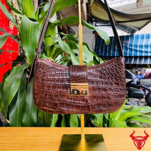 Hình ảnh túi đeo vai nữ da bụng cá sấu HM-19 màu nâu, khóa vàng, đặt trên giá gỗ giữa cây xanh và chợ, nổi bật phong cách sang trọng.