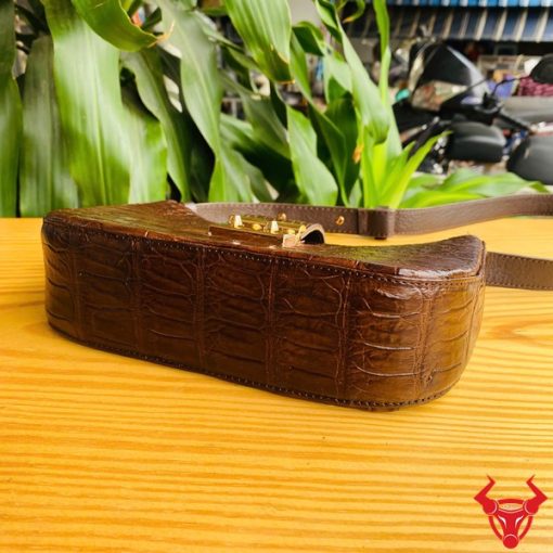 Hình ảnh túi đeo vai da bụng cá sấu HM-19 màu nâu, khóa vàng, đặt trên giá gỗ, thể hiện phong cách sang trọng và cá tính.