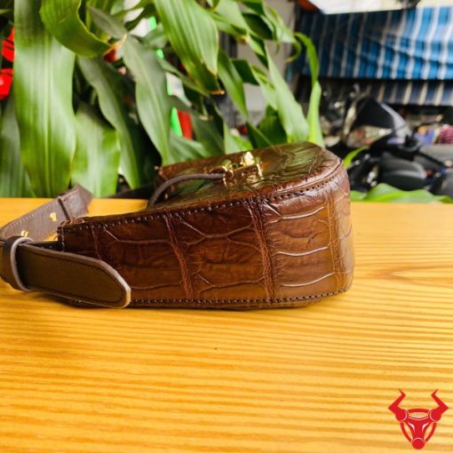 Hình ảnh túi đeo vai da bụng cá sấu HM-19 màu nâu, khóa vàng, đặt trên giá gỗ, thể hiện phong cách thời trang sang trọng và cá tính.