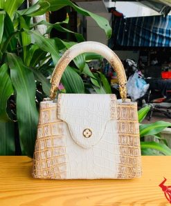 Hình ảnh túi xách nữ da cá sấu bạch tạng nhũ vàng, đặt trên bàn gỗ với nền cây xanh và chợ, thể hiện sự sang trọng.