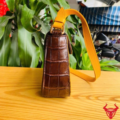 Hình ảnh túi xách da bụng cá sấu HM-18, nâu vàng viền da bò, đặt trên giá gỗ, nổi bật với phong cách sang trọng.