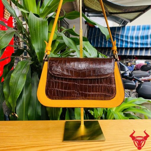 Hình ảnh túi xách nữ da bụng cá sấu HM-18, màu nâu vàng, viền da bò, đặt trên giá gỗ giữa cây xanh và chợ, nổi bật phong cách sang trọng.