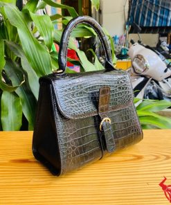 Hình ảnh túi xách HM-23 da cá sấu màu đen, kiểu dáng quý phái, khóa vàng, trên bàn gỗ với lá xanh và xe máy phía sau.