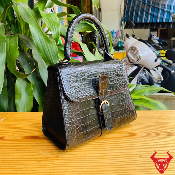 Thời trang sang trọng HM-23 cho phái đẹp Hình ảnh túi xách HM-23 da cá sấu màu đen, kiểu dáng quý phái, khóa vàng, trên bàn gỗ với lá xanh và xe máy phía sau.