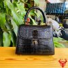 Hình ảnh túi xách da cá sấu HM-23 màu đen, thiết kế quý phái, khóa vàng, đặt trên bàn gỗ với nền lá xanh và xe máy mờ phía sau.