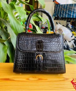 Hình ảnh túi xách da cá sấu HM-23 màu đen, thiết kế quý phái, khóa vàng, đặt trên bàn gỗ với nền lá xanh và xe máy mờ phía sau.