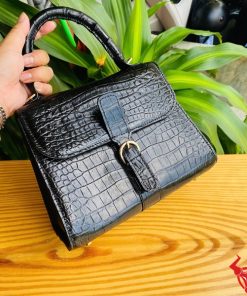 Hình ảnh túi xách HM-23 da cá sấu màu đen, kiểu dáng quý phái, khóa vàng nổi bật, trên bàn gỗ với lá xanh và xe máy phía sau.
