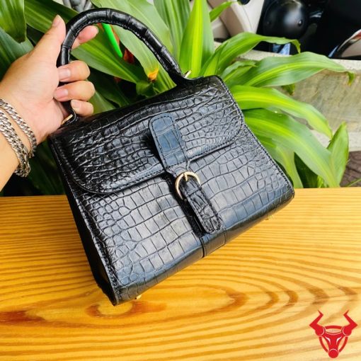Hình ảnh túi xách HM-23 da cá sấu màu đen, kiểu dáng quý phái, khóa vàng nổi bật, trên bàn gỗ với lá xanh và xe máy phía sau.