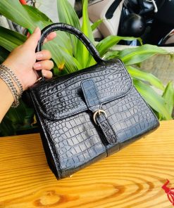 Hình ảnh túi xách da cá sấu HM-23 màu đen, thiết kế sang trọng, khóa vàng, đặt trên bàn gỗ với lá xanh và xe máy mờ phía sau.