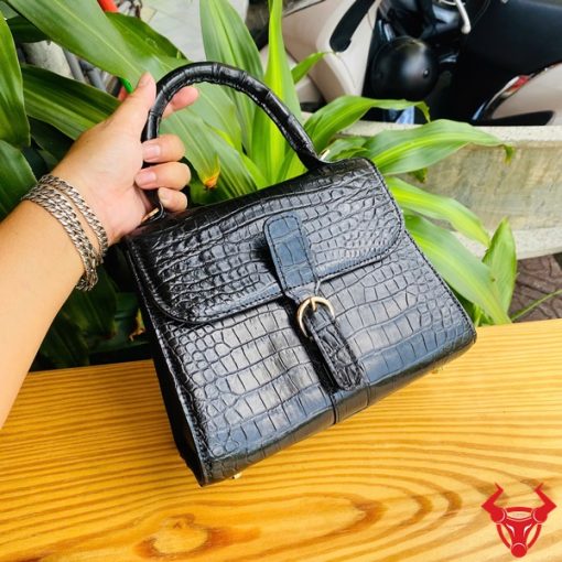 Hình ảnh túi xách da cá sấu HM-23 màu đen, thiết kế sang trọng, khóa vàng, đặt trên bàn gỗ với lá xanh và xe máy mờ phía sau.
