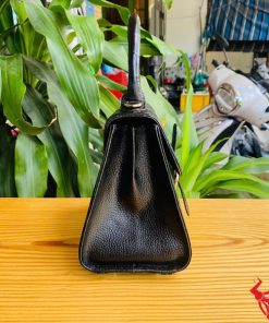 Hình ảnh túi xách da cá sấu HM-23 màu đen, kiểu dáng quý phái, khóa vàng, đặt trên bàn gỗ với lá xanh và xe máy mờ phía sau.
