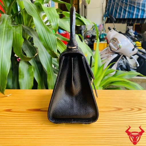 Hình ảnh túi xách da cá sấu HM-23 màu đen, kiểu dáng quý phái, khóa vàng, đặt trên bàn gỗ với lá xanh và xe máy mờ phía sau.