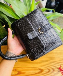 Hình ảnh túi xách HM-23 da cá sấu màu đen, thiết kế sang trọng, khóa vàng nổi bật, trên bàn gỗ với lá xanh và xe máy phía sau.