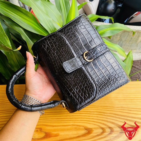 Túi xách quý phái HM-23 - Phong cách thời thượng Hình ảnh túi xách HM-23 da cá sấu màu đen, thiết kế sang trọng, khóa vàng nổi bật, trên bàn gỗ với lá xanh và xe máy phía sau.