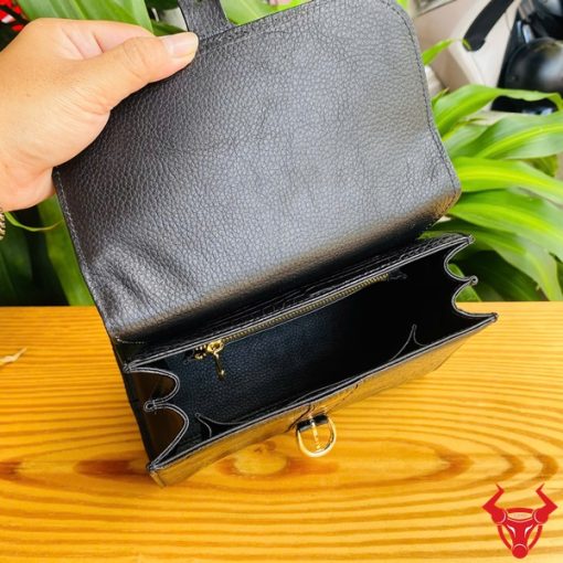 Hình ảnh túi xách HM-23 da cá sấu màu đen, thiết kế sang trọng, khóa vàng lấp lánh, trên bàn gỗ với lá xanh và xe máy phía sau.