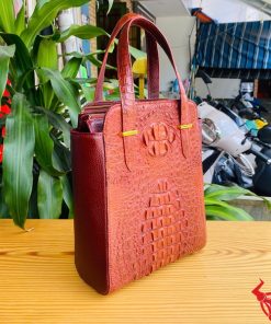 Hình ảnh túi xách HM-21 da cá sấu màu nâu đỏ, kiểu dáng cao cấp, khóa vàng, trên bàn gỗ với lá xanh và xe máy phía sau.