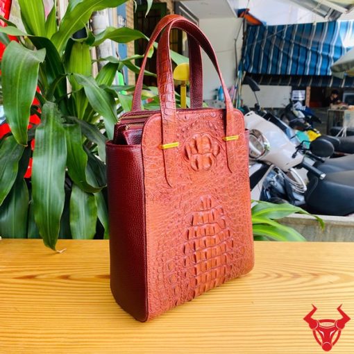 Hình ảnh túi xách HM-21 da cá sấu màu nâu đỏ, kiểu dáng cao cấp, khóa vàng, trên bàn gỗ với lá xanh và xe máy phía sau.