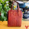 Hình ảnh túi xách da cá sấu HM-21 màu nâu đỏ, thiết kế cao cấp, khóa vàng, đặt trên bàn gỗ với nền lá xanh và xe máy mờ phía sau.