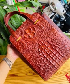 Hình ảnh túi xách da cá sấu HM-21 màu nâu đỏ, thiết kế cao cấp, khóa vàng, đặt trên bàn gỗ với lá xanh và xe máy mờ phía sau.