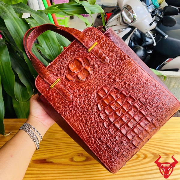 Vẻ đẹp sành điệu với túi xách HM-21 da cá sấu Hình ảnh túi xách da cá sấu HM-21 màu nâu đỏ, thiết kế cao cấp, khóa vàng, đặt trên bàn gỗ với lá xanh và xe máy mờ phía sau.