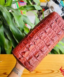 Hình ảnh túi xách HM-21 da cá sấu màu nâu đỏ, thiết kế cao cấp, khóa vàng, trên bàn gỗ với lá xanh và xe máy phía sau.