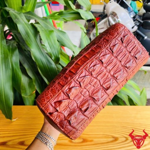 Hình ảnh túi xách HM-21 da cá sấu màu nâu đỏ, thiết kế cao cấp, khóa vàng, trên bàn gỗ với lá xanh và xe máy phía sau.