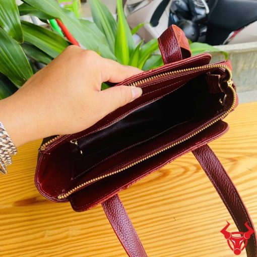 Hình ảnh túi xách da cá sấu HM-21 màu nâu đỏ, kiểu dáng cao cấp, khóa vàng, đặt trên bàn gỗ với lá xanh và xe máy mờ phía sau.
