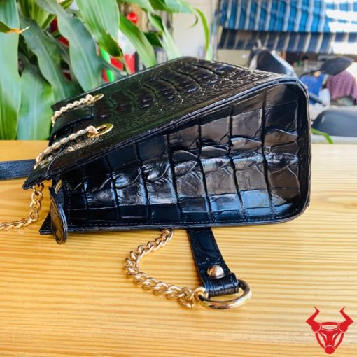 Hình ảnh túi đeo chéo da cá sấu gù TARA4 màu đen, khóa vàng, dây xích, đặt trên bàn gỗ với lá xanh và xe máy mờ phía sau.