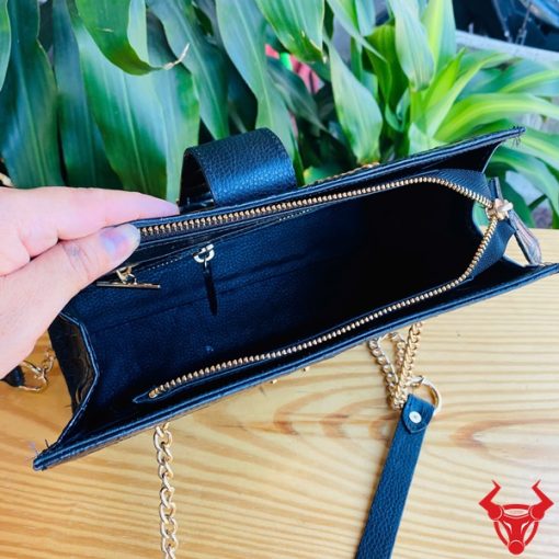Hình ảnh túi đeo chéo TARA4 da cá sấu gù màu đen, khóa vàng, dây xích, trên bàn gỗ với lá xanh và xe máy phía sau.
