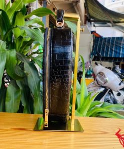 Hình ảnh túi xách da cá sấu HM-22 hình trái tim màu đen, thiết kế sang trọng, khóa vàng, đặt trên giá gỗ với lá xanh và xe máy mờ phía sau.