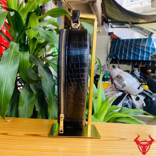 Hình ảnh túi xách da cá sấu HM-22 hình trái tim màu đen, thiết kế sang trọng, khóa vàng, đặt trên giá gỗ với lá xanh và xe máy mờ phía sau.