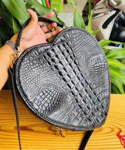 Hình ảnh túi xách da cá sấu HM-22 hình trái tim màu đen, thiết kế sang trọng, khóa vàng, đặt trên giá gỗ với lá xanh và xe máy mờ phía sau.