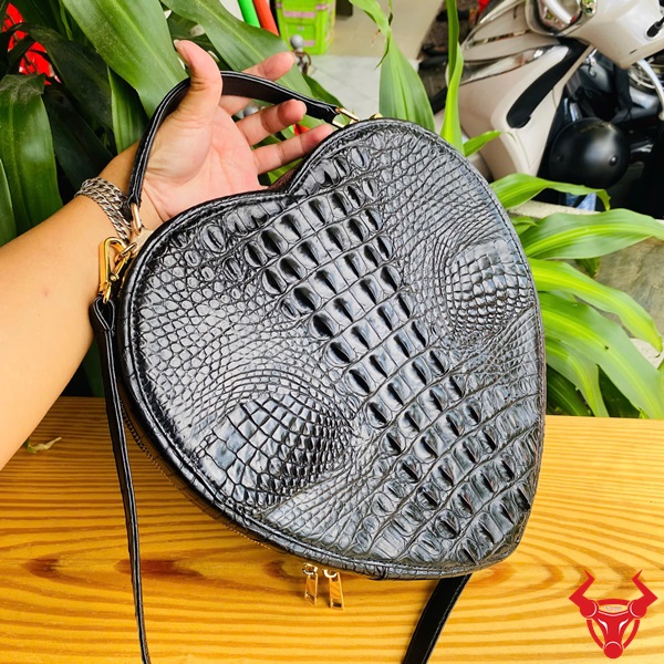 Vẻ đẹp sành điệu với túi xách HM-22 da cá sấu Hình ảnh túi xách da cá sấu HM-22 hình trái tim màu đen, thiết kế sang trọng, khóa vàng, đặt trên giá gỗ với lá xanh và xe máy mờ phía sau.