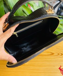 Hình ảnh túi xách da cá sấu HM-22 hình trái tim màu đen, kiểu dáng sang trọng, khóa vàng, đặt trên giá gỗ với lá xanh và xe máy mờ phía sau.