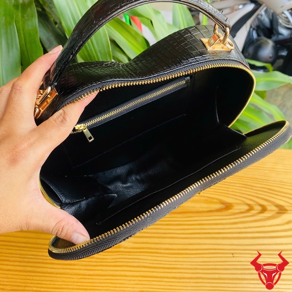 Thời trang sang trọng HM-22 cho phái đẹp Hình ảnh túi xách da cá sấu HM-22 hình trái tim màu đen, kiểu dáng sang trọng, khóa vàng, đặt trên giá gỗ với lá xanh và xe máy mờ phía sau.