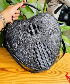 Hình ảnh túi xách HM-22 da cá sấu hình trái tim màu đen, thiết kế quý phái, khóa vàng nổi bật, trên giá gỗ với lá xanh và xe máy phía sau.