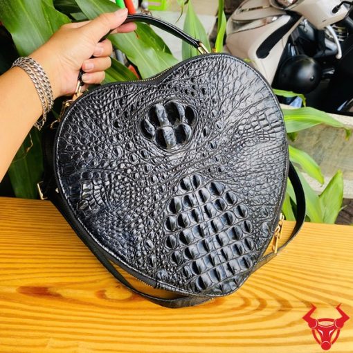 Hình ảnh túi xách HM-22 da cá sấu hình trái tim màu đen, thiết kế quý phái, khóa vàng nổi bật, trên giá gỗ với lá xanh và xe máy phía sau.