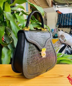 Hình ảnh túi xách da cá sấu HM-24 màu đen, kiểu dáng quý phái, khóa vàng lấp lánh, đặt trên bàn gỗ với lá xanh và xe máy phía sau.