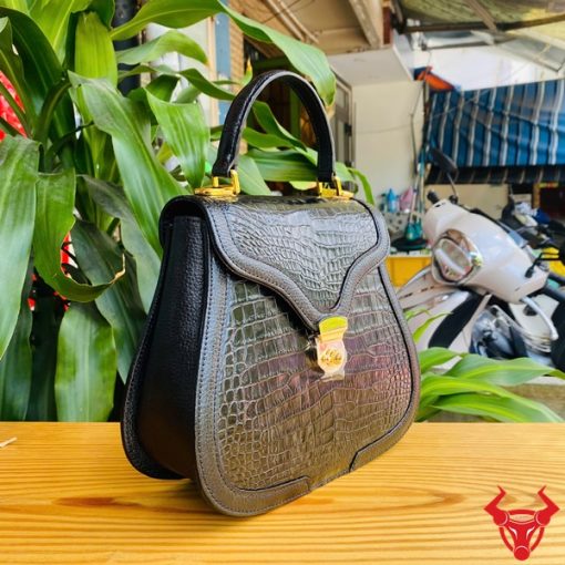 Hình ảnh túi xách da cá sấu HM-24 màu đen, kiểu dáng quý phái, khóa vàng lấp lánh, đặt trên bàn gỗ với lá xanh và xe máy phía sau.